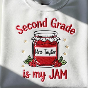 May include: White sweatshirt with red embroidered text that reads "Second Grade is my JAM" with a jar of jam and strawberries. The jar has a label that says "Mrs Taylor". Gold star accents are also embroidered.