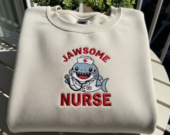 Pode incluir: Camisola creme com o texto bordado "JAWSOME NURSE". O desenho apresenta um tubarão de desenho animado usando um chapéu de enfermeira e um estetoscópio. O texto e o gráfico são em vermelho e preto.