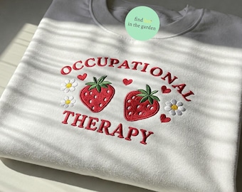 May include: White sweatshirt with red embroidered text that reads "OCCUPATIONAL THERAPY" with two red embroidered strawberries and white embroidered flowers.