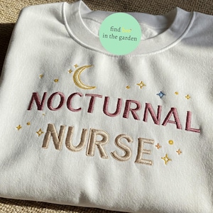 Op de afbeelding: Witte trui met geborduurde tekst "Nocturnal Nurse" in roze en goud. Het ontwerp bevat ook een halve maan en sterren.