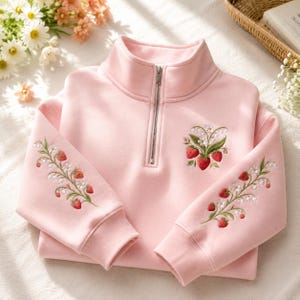 May include: A light pink quarter-zip sweatshirt featuring embroidered designs of red strawberries and white lily of the valley flowers on the chest and along the length of both sleeves. The garment has a high collar and ribbed cuffs.