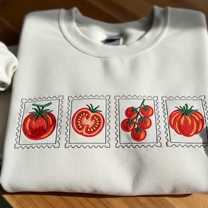Puede incluir: Sudadera color crema con un diseño bordado de cuatro sellos postales de tomate. Cada sello muestra una variedad diferente de tomate en tonos rojos y verdes, con detalles intrincados. La sudadera está hecha de un material suave y cómodo.