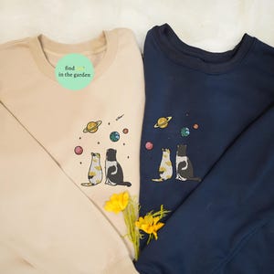 Puede incluir: Dos sudaderas con un diseño de temática espacial con dos gatos mirando hacia arriba a los planetas. El texto "find in the garden" está en la sudadera beige.