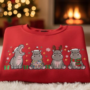 May include: A folded red sweatshirt with an embroidered design of four cartoon hippos. Each hippo is wearing festive holiday attire, including Santa hats, reindeer antlers, and a scarf. The design includes snowflakes, stars, and wrapped gifts.