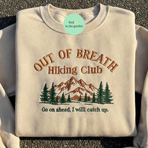 May include: A beige sweatshirt with the text "Out of Breath Hiking Club" embroidered in brown. The design also includes a green mountain range with trees and the text "Go on ahead, I will catch up."