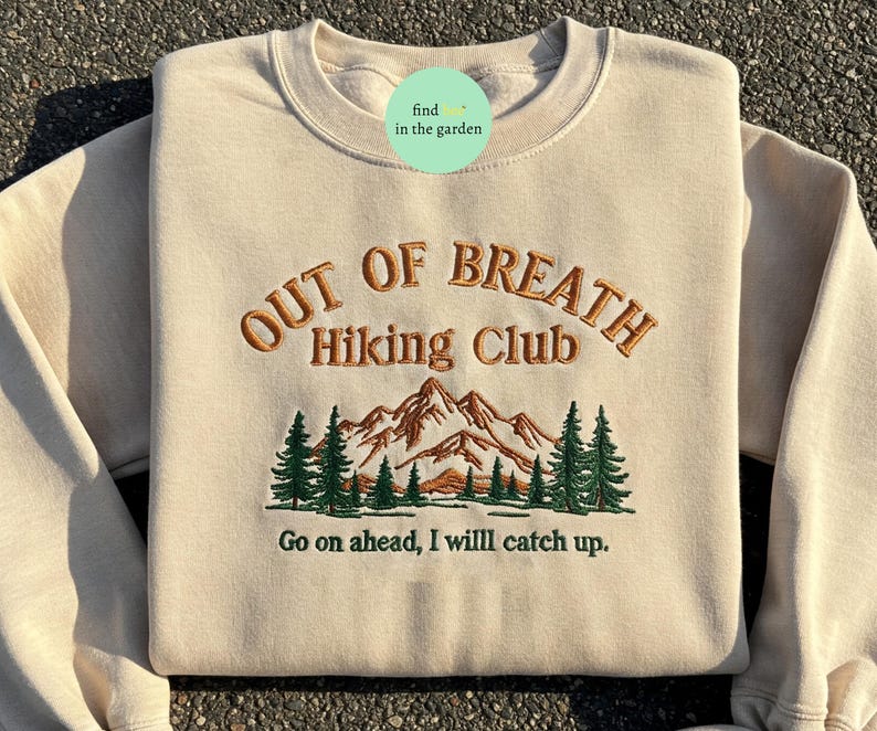 May include: A beige sweatshirt with the text "Out of Breath Hiking Club" embroidered in brown. The design also includes a green mountain range with trees and the text "Go on ahead, I will catch up."