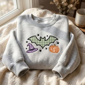 Puede incluir: Una sudadera gris claro de cuello redondo con un diseño bordado de un murciélago de cuadros verdes, un sombrero de bruja de cuadros morados y una calabaza de cuadros naranjas, rodeados de pequeñas estrellas negras bordadas en el frente.