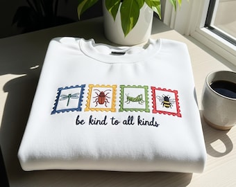 May include: White sweatshirt with embroidered insect postage stamp designs. The stamps feature a dragonfly, beetle, grasshopper, and bee. The phrase "be kind to all kinds" is embroidered below the stamps.