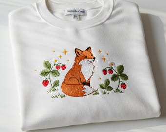 Pode incluir: Camisola branca com um desenho bordado com uma raposa, morangos e estrelas. A raposa é laranja e branca, sentada entre plantas de morango verdes com bagas vermelhas. O texto "Sample Count" está na etiqueta.