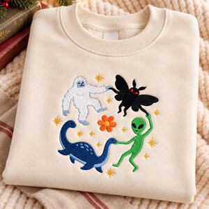 May include: A cream-coloured crewneck sweatshirt featuring an embroidered design of a white yeti, a black moth with red eyes, a blue long-necked dinosaur, and a green alien holding hands in a circle around a central orange flower.