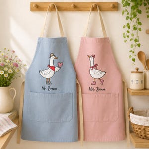 May include: Two aprons with beige neck straps and waist ties. The left apron is light blue with a graphic of a goose wearing a hat, scarf, and boots, holding a heart, labelled Mr. Brown. The right apron is pink with a goose wearing a bow, labelled Mrs. Brown.