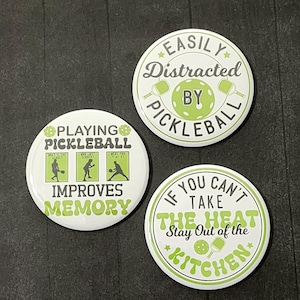 May include: Three round white buttons with green text and images. The first button says "Easily Distracted by Pickleball". The second button says "Playing Pickleball Improves Memory" and has three images of people playing pickleball. The third button says "If You Can't Take the Heat Stay Out of the Kitchen" and has a pickleball paddle and ball.