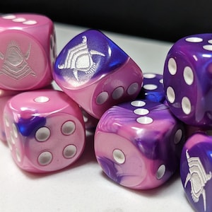 Puede incluir: Un conjunto de dados de seis caras en tonos morados y rosas con un acabado jaspeado. Algunos dados presentan puntos blancos, mientras que otros muestran un icono de casco grabado en blanco en una cara. Los dados están dispuestos en grupo.