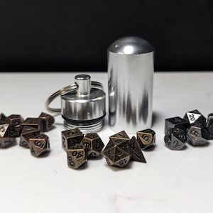 Può includere: Un set di piccoli dadi metallici a più facce con finiture color bronzo e grigio scuro, disposti in tre gruppi. Un contenitore cilindrico in metallo argentato con coperchio a vite e un portachiavi attaccato si trova al centro.