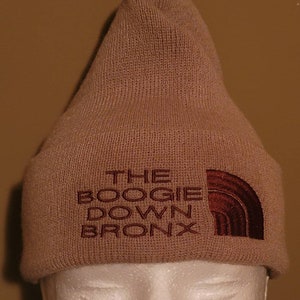 Puede incluir: Gorro de punto beige con un logotipo bordado en marrón que dice "THE BOOGIE DOWN BRONX".