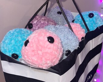 May include: A black and white striped shopping bag filled with several plush, round, colorful toys. The toys are in shades of blue, pink, and purple, each with two black button eyes. The bag has black handles.