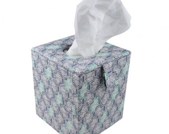 May include: A square tissue box cover featuring a geometric pattern of blue, teal, and white interlocking shapes. The cover has a top opening with a single white tissue pulled through, and a small fabric tab detail on the front side panel.