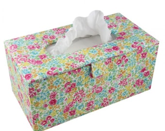 May include: A rectangular tissue box cover featuring a dense floral pattern with small pink, yellow, and light blue flowers on a white background. A single white tissue is pulled through the oval-shaped opening on the top of the box.