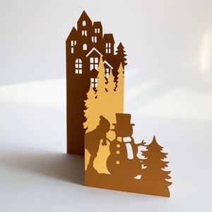 Puede incluir: Una tarjeta de papel marrón recortada con una escena de invierno. La tarjeta presenta una silueta de una casa con árboles y un muñeco de nieve. El muñeco de nieve lleva un sombrero y una bufanda y tiene una nariz de zanahoria. La tarjeta está doblada por la mitad y el interior está en blanco.