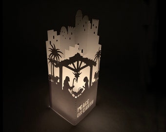 Può includere: Decorazione natalizia in carta bianca con una silhouette della scena della Natività, tra cui Maria, Giuseppe e Gesù bambino. La decorazione ha una città sullo sfondo e la scritta "Peace on Earth".