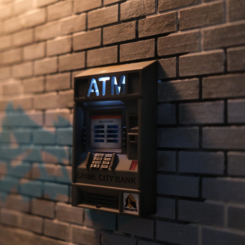 Puede incluir: Un cajero autom&aacute;tico gris con las palabras "GRIME CITY BANK" y "ATM" en la parte delantera. El cajero est&aacute; montado en una pared de ladrillo.