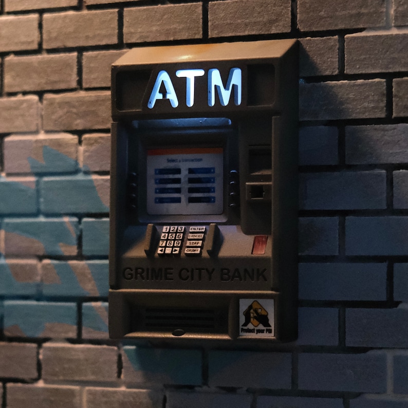 Puede incluir: Un cajero autom&aacute;tico gris con las palabras "GRIME CITY BANK" en la parte delantera. El cajero autom&aacute;tico tiene un teclado con n&uacute;meros y una pantalla que dice "Select a Transaction". El cajero autom&aacute;tico est&aacute; iluminado con una luz azul.