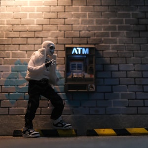 Puede incluir: Una figura blanca con una cara de calavera se encuentra frente a un cajero autom&aacute;tico con la palabra "ATM" impresa en &eacute;l. La figura lleva una chaqueta blanca, pantalones negros y zapatos negros. El cajero autom&aacute;tico est&aacute; contra una pared de ladrillo.