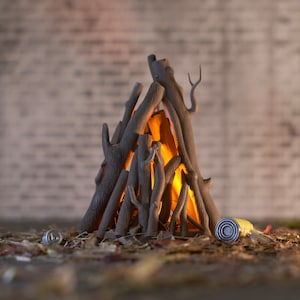 Puede incluir: Una hoguera de madera en miniatura con una luz naranja incandescente en su interior. La hoguera está hecha de pequeñas ramas marrones y está sentada sobre un lecho de hojas marrones.