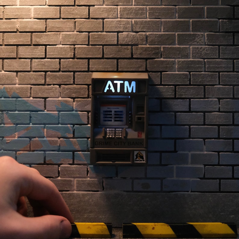 Puede incluir: Un cajero autom&aacute;tico gris con las palabras "GRIME CITY BANK" y "ATM" en &eacute;l, montado en una pared de ladrillo.