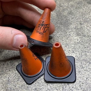 Könnte beinhalten: Miniatur-Leitkegel in Orange mit schwarzen Basen. Auf jedem Kegel steht das Wort "HATER" in schwarzer Graffiti-Schrift. Diese kleinen Kegel sind wahrscheinlich für ein Modell oder Dekorationszwecke gedacht.