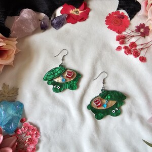 May include: A pair of dangle earrings shaped like green turtles with hinged shells that open to reveal a sandy interior featuring a miniature red cup and a blue shovel. The earrings are crafted from acrylic and feature silver-toned metal hooks.