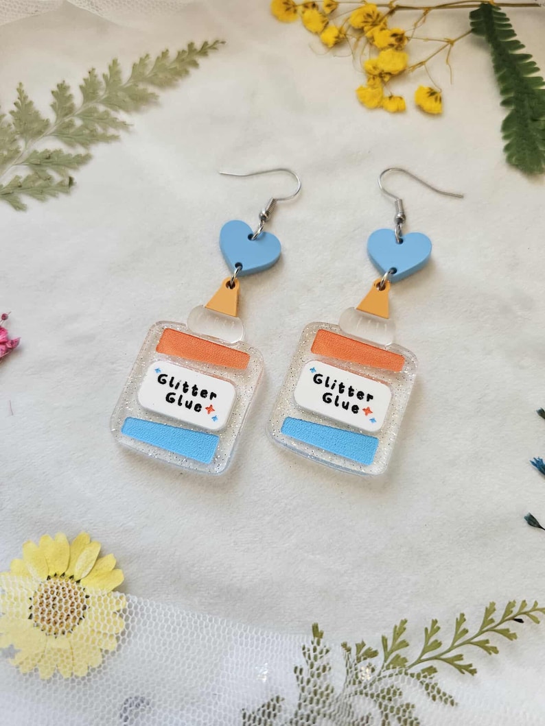 May include: A pair of novelty earrings featuring a clear, glittery glue bottle design with orange and blue accents. Each earring has a blue heart-shaped top and the words "Glitter Glue+" printed on the label. The earrings hang from silver-colored hooks.