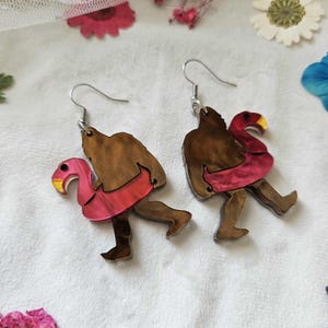 May include: A pair of novelty earrings featuring a brown figure with a pink flamingo float. The earrings have silver-colored hooks. The flamingo float is pink with a yellow beak. The figure is brown with a red inner tube.