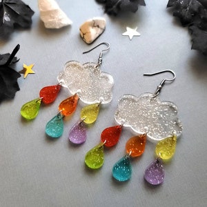 May include: A pair of rainbow-colored raindrop earrings with a glitter cloud charm. The earrings are made of clear acrylic and have a silver hook.