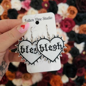 May include: A pair of silver heart-shaped earrings with black glitter and the word "Blesgh" written in gothic font. The earrings are on a white card with the text "Fallen Pine Studio".