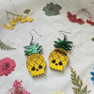 May include: A pair of yellow and black pineapple skull earrings with green tops. The earrings have a skull design with crosshatch detailing and are suspended from silver-colored hooks. The earrings are set against a white background with dried flowers.