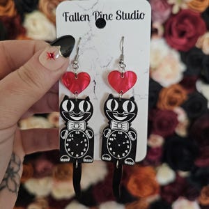 May include: Black and white cat earrings with red heart accents. The earrings feature a clock face on the cat's belly and a dangling tail. The earrings are attached to a white card with the text "Fallen Pine Studio".