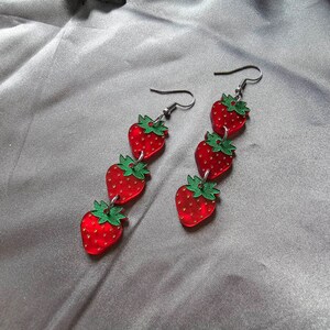 May include: A pair of dangle earrings featuring four red and green strawberry charms hanging from silver hooks.