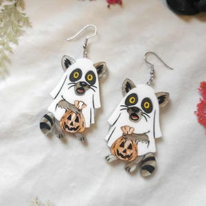 May include: A pair of Halloween-themed earrings featuring raccoon characters dressed as ghosts. Each white ghost-like figure has a raccoon face with yellow eyes and is holding a carved pumpkin. The earrings have silver-colored hooks.