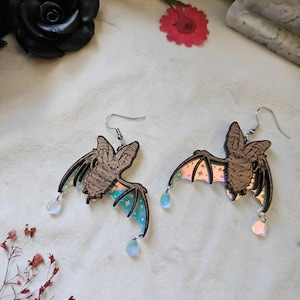 May include: A pair of bat-shaped earrings. The earrings feature a brown bat design with iridescent wings and a small, clear teardrop charm. The wings have a blue and green color with a star pattern. The earrings hang from silver-colored hooks.