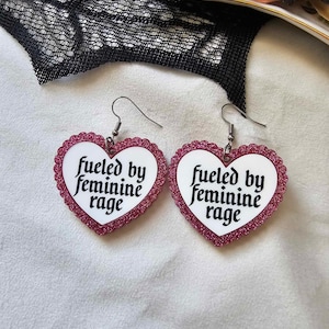 May include: A pair of pink glitter heart-shaped earrings with the text "fueled by feminine rage" on each earring.