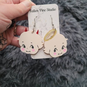 May include: A pair of earrings featuring a white cherub with a halo and a white cherub with horns. The earrings are on a card that says "Fallen Pine Studio".