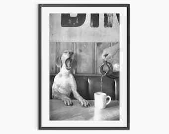May include: A black and white framed wall art print featuring a dog with its mouth wide open sitting at a table. A hand pours liquid from a glass carafe into a white ceramic mug. The background shows a booth with vertical paneling and text.