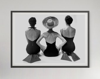 May include: Three women in black swimsuits sit on red and white beach mats, facing away from the camera. The woman in the middle is wearing a large, wide-brimmed straw hat.