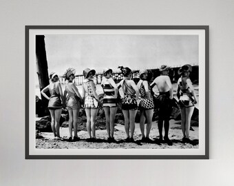 May include: A black and white framed photograph depicting eight individuals standing in a row on a sandy beach, wearing vintage-style swimsuits with various patterns including stripes, polka dots, and checks, all facing away from the camera.