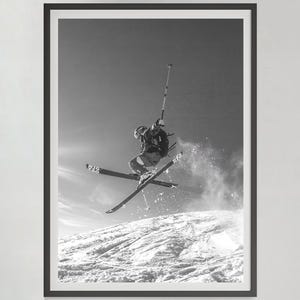 May include: Black and white framed print of a skier mid-air, performing a jump on a snowy slope. The skier is wearing a helmet and winter gear, with ski poles in hand. The background shows a bright sky.