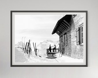 May include: A black and white framed wall art print depicting a mountain lodge with a balcony. Two figures stand on the balcony overlooking snow-covered peaks. Several pairs of vintage wooden skis are stuck upright in the snow in the foreground.
