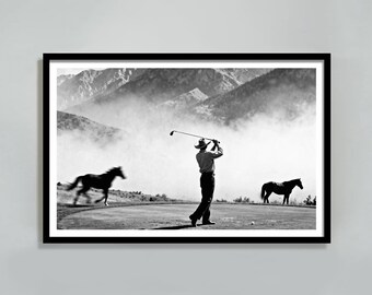 May include: A black and white framed print depicting a person wearing a cowboy hat swinging a golf club on a course. A horse is captured in motion on the left, while another stands still on the right. Mountains shrouded in mist appear in the background.