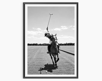 May include: A black and white photograph of a polo player riding a horse on a grass field. The player wears a helmet and boots, holding a mallet high in the air while galloping toward the camera. A polo ball is visible on the ground in front.