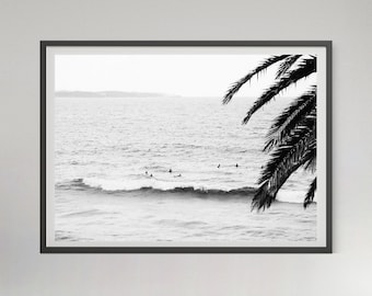 May include: A black and white framed wall art print featuring an ocean scene with surfers waiting for waves in the water. A palm tree frond hangs in the upper right corner, partially obscuring the view of the horizon and the sea.